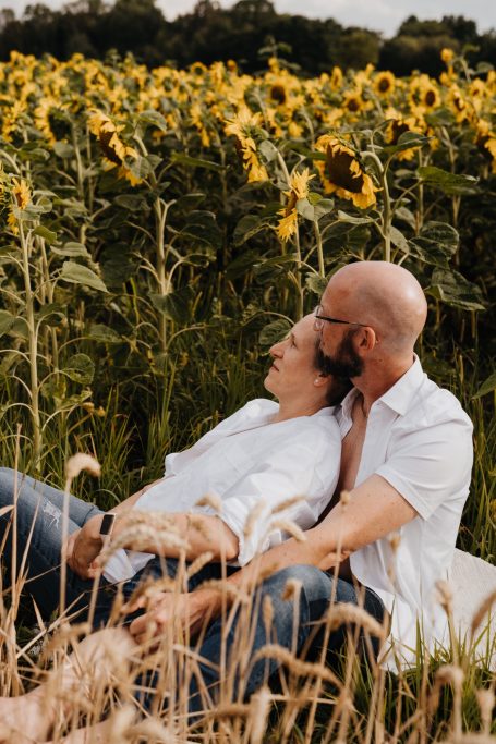 Paar sitzt in einem Sonnenblumenfeld, entspannt und liebevoll umarmt.
