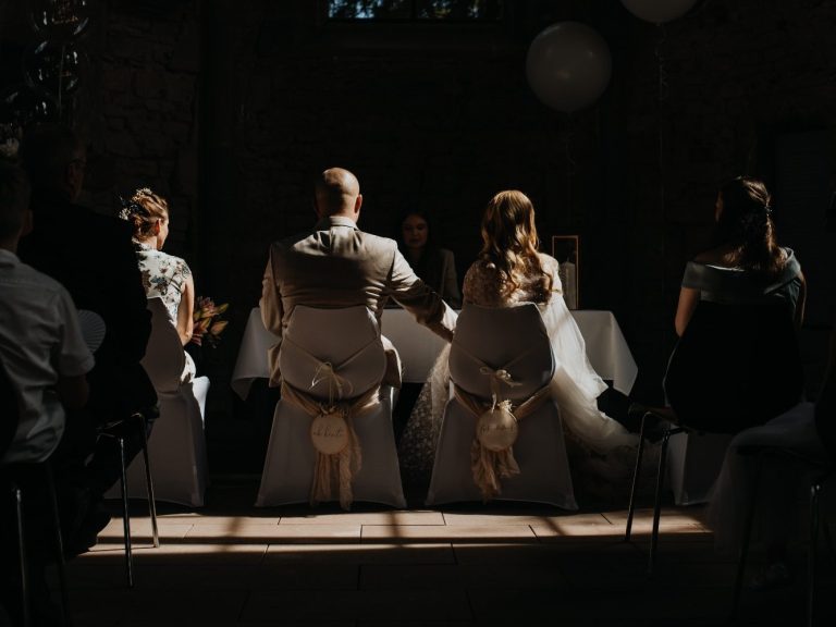 Brautpaar und Gäste sitzen in einem festlichen Raum, Licht fällt sanft auf die Szene.