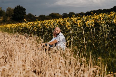 Mann sitzt im hohen Gras vor einem Sonnenblumenfeld unterbewölktem Himmel.