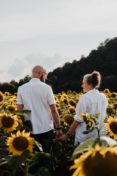 Paar hält Händchen in einem Sonnenblumenfeld unter einem bewölkten Himmel.